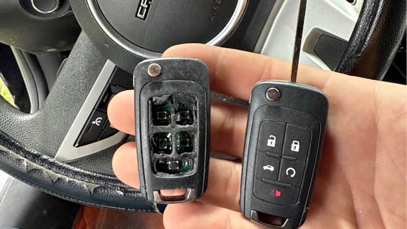 A worn GM flip key with its button pad worn through, held next to a replacement flip key in front of a Camaro steering wheel